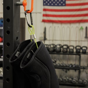 May include: Two black knee pads with a green strap hanging from a silver carabiner attached to an orange hook on a black metal rack. The rack has a label that reads "INFINITY" and "MADE IN THE U.S.A."