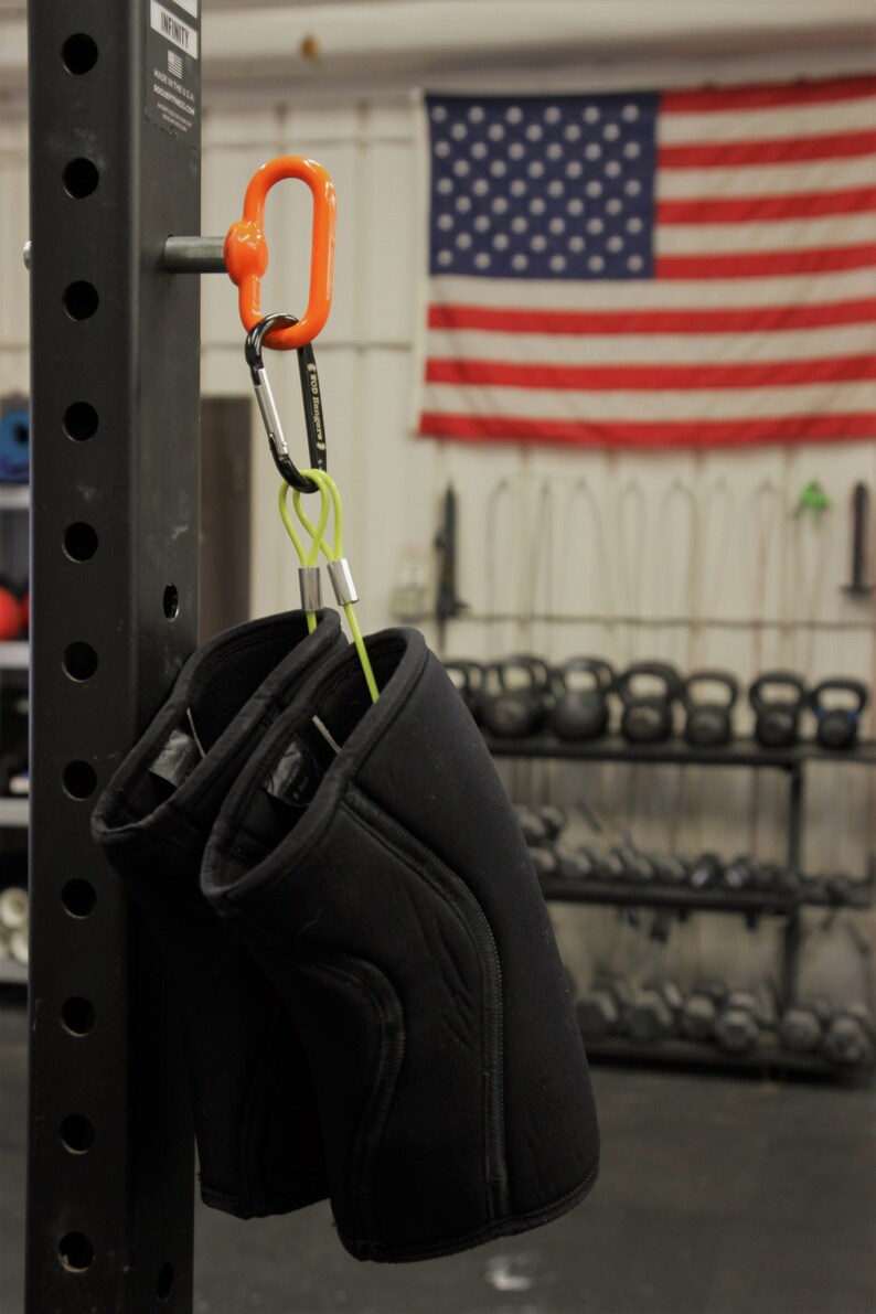 May include: Two black knee pads with a green strap hanging from a silver carabiner attached to an orange hook on a black metal rack. The rack has a label that reads "INFINITY" and "MADE IN THE U.S.A."