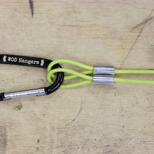 May include: A black carabiner with the text "WOD Hangers" on it is attached to two lime green ropes with silver metal sleeves.