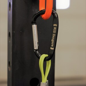 May include: A black carabiner with the text "WOD Hangers" engraved on the side. The carabiner is attached to a bright orange loop and a lime green cord with a silver metal clip.
