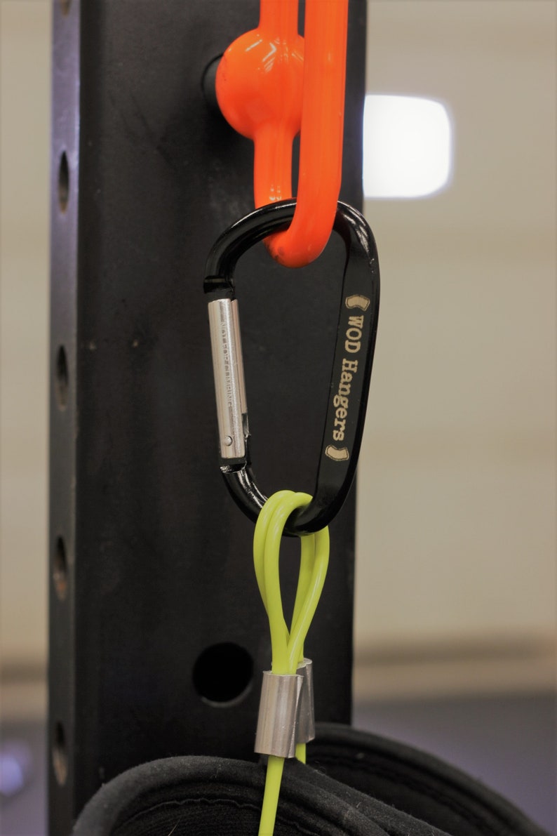 May include: A black carabiner with the text "WOD Hangers" engraved on the side. The carabiner is attached to a bright orange loop and a lime green cord with a silver metal clip.