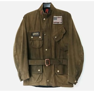 May include: An olive green Barbour International jacket featuring a stand-up collar, a belted waist, and four front pockets with snap closures. The chest displays a patch with an American flag and the text American Team I.S.D.T. 1964.