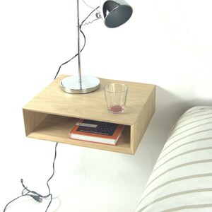 Op de afbeelding: Een zwevend nachtkastje van licht hout met een plank, met een boek en een glas. Boven het nachtkastje is een zilveren bureaulamp gemonteerd. Het bed heeft een gestreept dekbedovertrek. De tafel is ongeveer 40 cm breed.