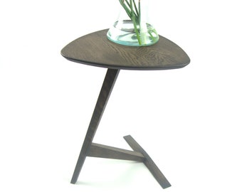 May include: A dark brown, triangular side table with a single leg and a three-legged base. A clear glass vase with yellow flowers sits on top. The table is made of wood and has a modern design.
