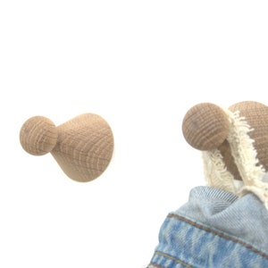 Peut inclure: Deux crochets muraux en bois avec un bouton arrondi sur le dessus et une base en forme de cône. Un crochet a une veste en jean bleue accrochée.