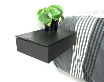 May include: A black floating bedside table with a rectangular wooden drawer and a flat top surface. A small green plant in a black pot sits on the bedside table, which is mounted next to a bed with a grey and white striped comforter.