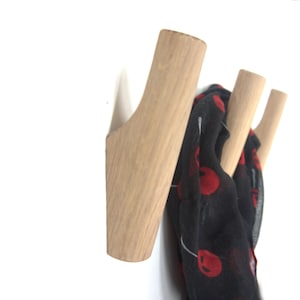 Può includere: Appendiabiti da parete in legno con tre ganci. I ganci sono di colore marrone chiaro e sostengono una sciarpa nera con motivi circolari rossi. L'appendiabiti è montato su una parete bianca.