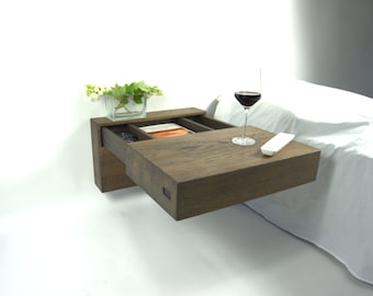 Könnte beinhalten: Ein schwebender Nachttisch aus dunklem Holz mit einer offenen Schublade, in der sich Gegenstände wie ein Buch und Büroklammern befinden. Ein Glas Rotwein und eine Fernbedienung stehen auf der Tischplatte. Eine kleine Glasvase mit Grünzeug steht auf dem Nachttisch.