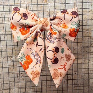 Può includere: Un grande fiocco decorativo con sfondo color pesca, decorato con zucche, orecchie di Topolino e motivi autunnali. Il fiocco viene probabilmente utilizzato come accessorio per capelli o per confezioni regalo.