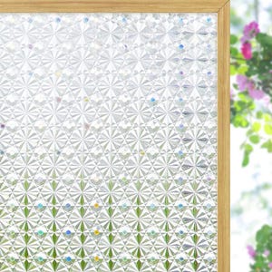 May include: A window film with a geometric pattern of silver and white shapes, creating a sparkling effect. The film is framed in light brown wood. The background shows green foliage and colorful dots.