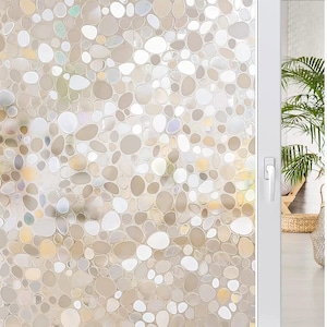 May include: Window film with a pebble design. The film is composed of various sized, rounded shapes in shades of white, beige, and tan. The film is installed on a sliding glass door. A white door frame and a glimpse of a room are visible.