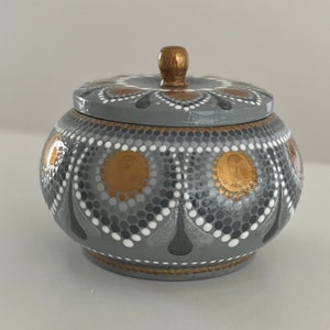 May include: A small gray and gold painted wooden box with a lid. The box is decorated with a pattern of dots and circles.