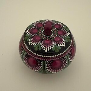May include: A hand-painted, round decorative jar with a lid. The jar is black with a pattern of pink and white dots, and green leaf-shaped designs. The lid has a small, round knob.