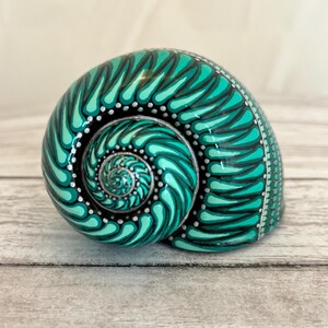 May include: A hand-painted seashell with a teal and black swirl pattern. The shell is decorated with white dots and silver accents.