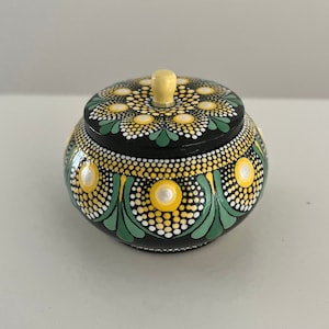 May include: A small, round, black wooden box with a lid. The box is decorated with yellow and green dots and a white knob on the lid.