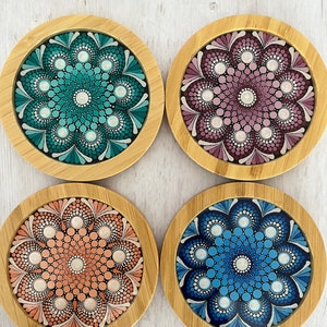 May include: Set of four round, wooden coasters with a mandala design. Each coaster features a different color scheme: teal, pink, peach, and blue. The coasters are perfect for protecting surfaces from heat and scratches.