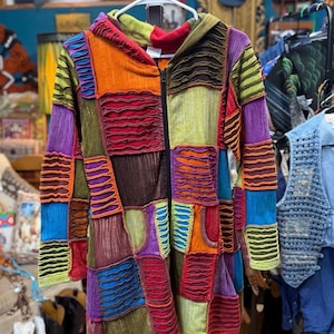 May include: A long-sleeve hooded jacket featuring a patchwork design of various rectangular fabric panels in shades of orange, green, purple, blue, and brown. The garment has a black front zipper and textured, ruffled fabric detailing.