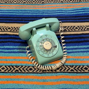 May include: A vintage turquoise rotary dial telephone with a coiled cord. The phone is set against a colorful striped textile background with blue, orange, and black patterns. The dial features numbers and letters.