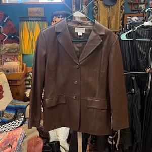 May include: A dark brown leather blazer featuring a notched lapel, three front buttons, and two flap pockets at the waist. The interior label displays the brand name Pendleton. The jacket has long sleeves and a structured, tailored fit.