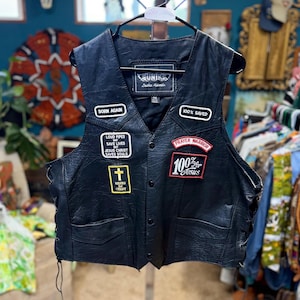 May include: A black leather vest featuring multiple embroidered patches including text that reads BORN AGAIN, 100% SAVED, LOUD PIPES MAY SAVE LIVES BUT JESUS CHRIST SAVES SOULS, PRAYER WARRIOR, 100% FOR JESUS, and a cross symbol with BIKERS FOR CHRIST.