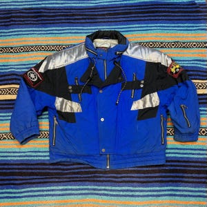 May include: A blue, black, and silver vintage ski jacket with a high collar and multiple zippers. The jacket features patches on the sleeves and shoulders, and a drawstring hood. The jacket is laid out on a striped textile.
