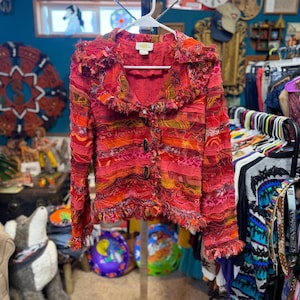 May include: A long-sleeve jacket in shades of red, orange, and yellow, featuring a textured, layered fabric construction with frayed edges, a wide collar, and two dark oval buttons down the front. The garment has a label reading Candy Canyon.