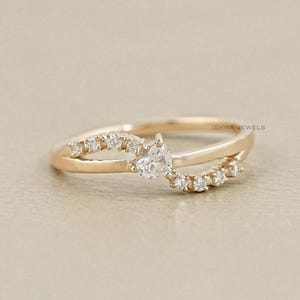 Op de afbeelding: Een goudkleurige ring met een gesplitst bandontwerp. De ring heeft een peervormige, heldere edelsteen in het midden, omringd door kleinere, ronde edelstenen. De band buigt om de vinger en creëert een delicate en elegante uitstraling.