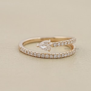 May include: A rose gold ring with a pear-shaped diamond at the centre. The ring features a split shank design, with small, round diamonds set along the bands. The ring is displayed on a neutral background.