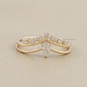 Könnte beinhalten: Ein goldfarbener Ring mit einem birnenförmigen Diamanten und kleineren runden Diamanten. Der Ring hat ein Doppelband-Design mit einer pfeilförmigen Spitze. Der Ring ist auf einem neutralen Hintergrund platziert.