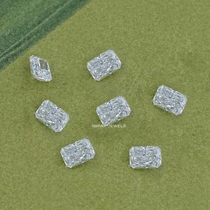 Op de afbeelding: Een verzameling kleine, rechthoekige, heldere edelstenen verspreid over een groene ondergrond. De edelstenen hebben een textuur, die het licht reflecteert. De achtergrond is effen, gedempt groen, wat een contrast vormt met de sprankelende edelstenen.