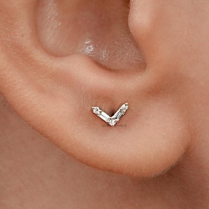 May include: A close-up of a person's ear with a small, silver, V-shaped earring with clear stones.