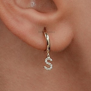 May include: A gold hoop earring with a dangling letter 'S' charm. The charm is encrusted with small, clear stones.