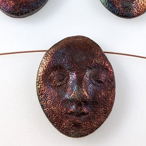 May include: Three iridescent brown ceramic beads with a face design. The beads are oval shaped and have a textured surface.