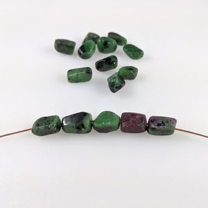 May include: A close-up of a wire strung with green and purple gemstone beads. The beads are irregular in shape and have a speckled appearance. There are additional loose beads in the background.