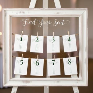 May include: White wooden frame with a glass front displaying a wedding seating chart. The chart is labeled "Find Your Seat" and has eight cards with numbers 1 through 8 hanging from twine with clothespins.