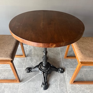 May include: A round wooden table with a dark brown finish and a thick edge, supported by a black cast iron pedestal base with four ornate feet, flanked by two chairs featuring light brown textured seats and wooden frames.
