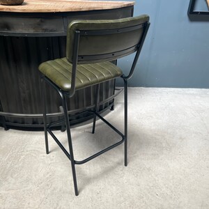 May include: A green leather bar stool with a black metal frame. The stool has a backrest and a footrest. The stool is in a room with a concrete floor.