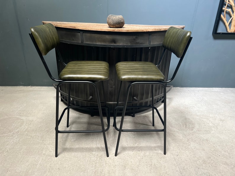 May include: Two green leather bar stools with black metal frames. The stools have a backrest and a footrest. The stools are in front of a black metal bar counter.