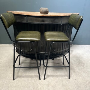 May include: Two green leather bar stools with black metal frames. The stools have a backrest and a footrest. The stools are in front of a black metal bar counter.