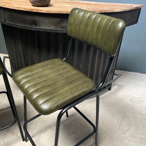 May include: Two green leather bar stools with black metal frames. The stools have a backrest and a seat with vertical stitching. The stools are in front of a black metal bar counter.