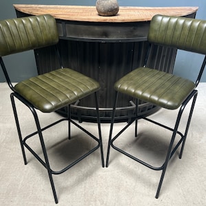 May include: Two green leather bar stools with black metal frames. The stools have a backrest and a footrest.