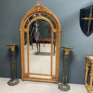 May include: A large, ornate gold-framed mirror with an arched top and intricate carvings. The mirror is flanked by two green marble pedestal stands.