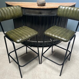 May include: Two green leather bar stools with black metal frames. The stools have a backrest and a footrest.