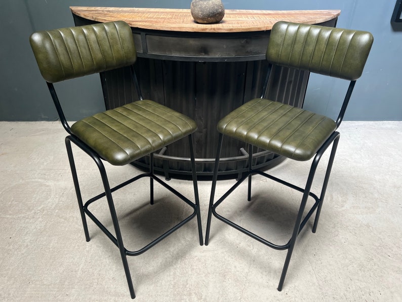 May include: Two green leather bar stools with black metal frames. The stools have a backrest and a footrest.
