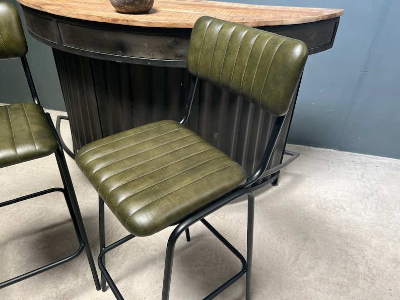 May include: Two green leather bar stools with black metal frames. The stools have a backrest and a seat with vertical stitching. The stools are in front of a black metal bar counter.