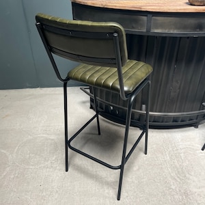 May include: A green leather bar stool with a black metal frame. The stool has a backrest and a footrest. The stool is in front of a black metal bar counter.