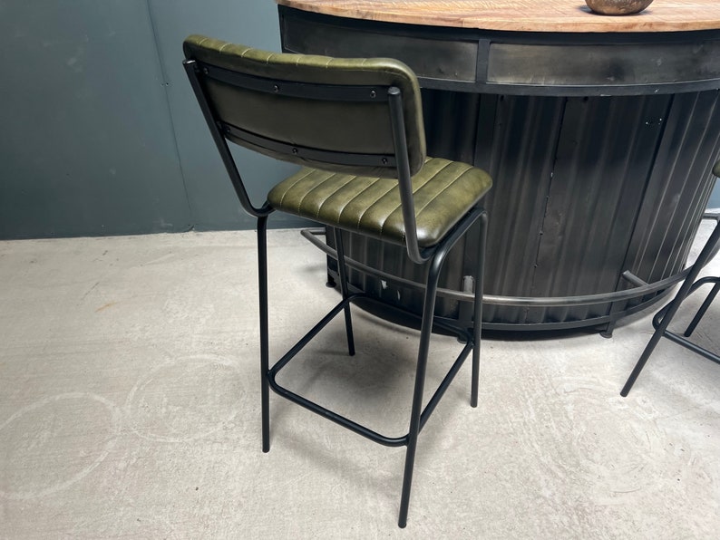May include: A green leather bar stool with a black metal frame. The stool has a backrest and a footrest. The stool is in front of a black metal bar counter.