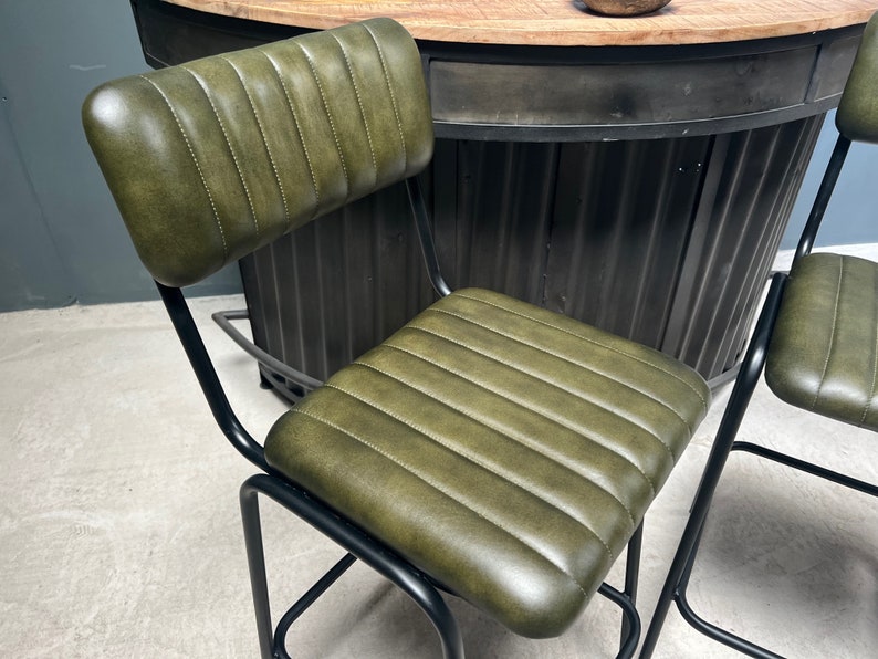 May include: Two green leather bar stools with black metal frames. The stools have a tall backrest and a padded seat. The stools are in front of a black metal bar counter.
