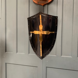 May include: A black metal shield with a gold cross design. The shield is hanging on a grey wall.