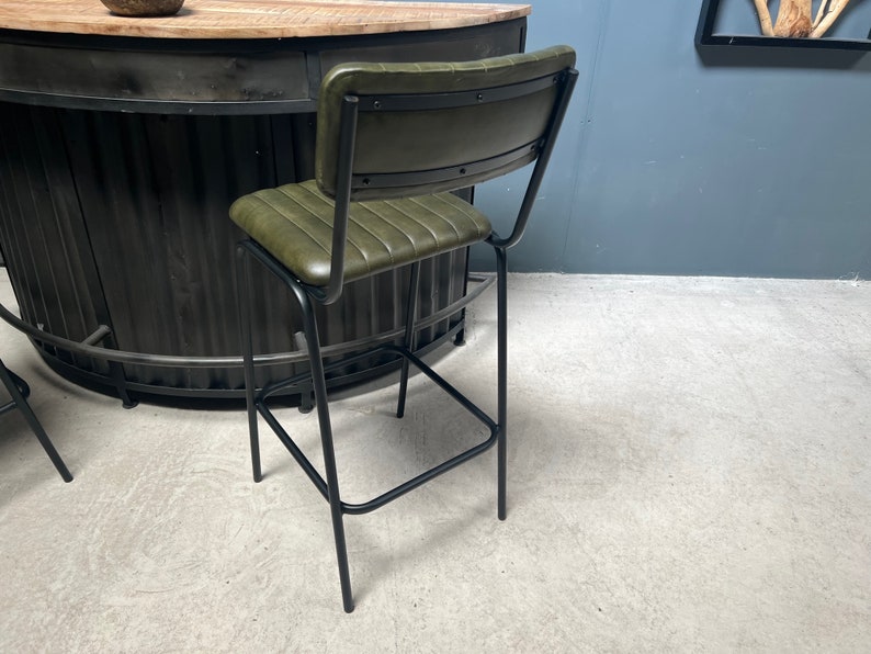 May include: A green leather bar stool with a black metal frame. The stool has a backrest and a footrest. The stool is in a room with a concrete floor.